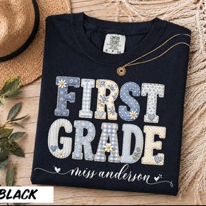 May include: A black t-shirt featuring the text FIRST GRADE in a patchwork style with blue, white, and cream floral and gingham patterns. Small daisies and hearts decorate the letters. Below, the text miss anderson is printed in white script.