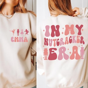 May include: Cream-colored sweatshirt with ballet-themed graphics and the name "Emma" in pink. The back features the phrase "In My Nutcracker Era" in shades of pink and red, with a nutcracker graphic.