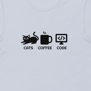 May include: Light blue t-shirt with a black graphic featuring a cat, coffee mug, and computer screen with code symbols. The words "CATS COFFEE CODE" are printed below the images. The shirt has short sleeves and a crew neck.