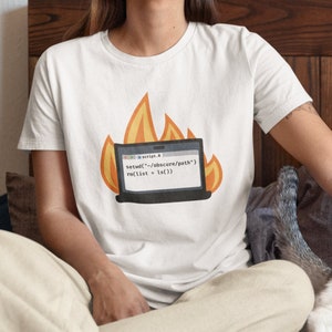 May include: A white t-shirt featuring a burning laptop graphic with code displayed. The person wearing the shirt is sitting on a bed with a cat. A teal and white logo is in the top left corner of the image.