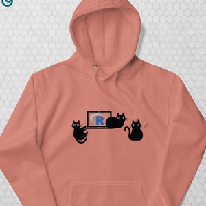 以下が含まれることがあります： フロントポケットとドローストリングフードが付いたコーラル色のパーカー。デザインには、3匹の黒い猫のシルエット、文字「R」を表示するラップトップ、左上に「the coding cats」というテキストが含まれています。