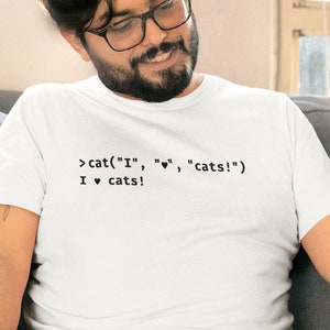 May include: White t-shirt with the text "cat("I", "♥", "cats!") I ♥ cats!" in black. The person is sitting on a grey sofa with a tabby cat. The cat has brown and white fur.