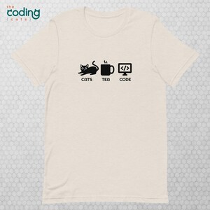 Op de afbeelding: Een wit T-shirt met een zwarte afbeelding van een kat, een kopje thee en een computerscherm met code op het scherm. De tekst onder de afbeelding luidt "Katten, Thee, Code."