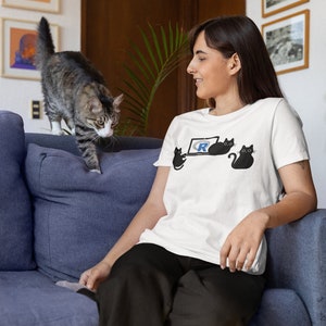 Puede incluir: Una mujer lleva una camiseta blanca con un gráfico de un gato negro. El gráfico muestra tres gatos, uno usando una computadora portátil con la letra "R" en la pantalla. La mujer está sentada en un sofá azul con un gato atigrado saltando en la parte posterior del sofá.