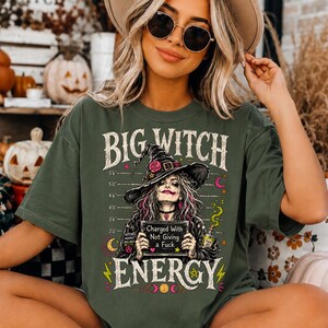 May include: An olive green t-shirt featuring a graphic of a witch in a mugshot lineup with height markers in feet. The text reads BIG WITCH ENERGY and Charged With Not Giving a Fuck. The design includes a potion bottle, stars, and moons.