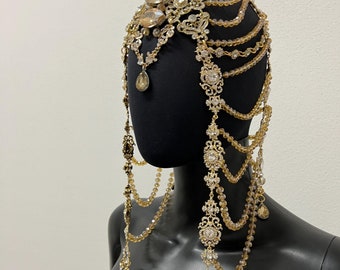 May include: A gold and crystal headpiece with dangling chains and teardrop-shaped crystals. The headpiece is on a black mannequin head.