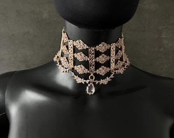 May include: A gold choker necklace with a delicate, intricate design featuring clear crystals. The necklace has a dangling crystal pendant.