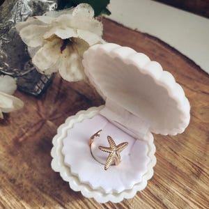 May include: A gold starfish ring presented in a white, shell-shaped jewellery box. The ring features a textured starfish design and sits on white fabric. The open jewellery box, displaying the ring, rests on a wooden surface.