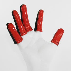 May include: A hand wearing white gloves with red finger caps. The finger caps are glossy and have a black trim. The gloves are against a white background.