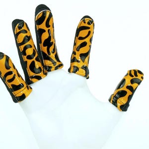 May include: Finger covers with a leopard print design. The covers are yellow with black spots and black trim. The covers are on a white hand.