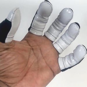 May include: Finger protectors with white, glossy caps and black accents are shown. The protectors are designed to fit over the fingers. The thumb protector has a black and white design. The protectors are shown on a hand.