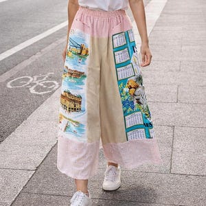 May include: A person wearing a white t-shirt and wide-leg trousers with a patchwork design. The trousers feature pink, beige, and patterned panels with images of buildings and calendars. They are wearing white trainers and walking on a city street.