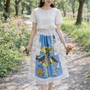 May include: A woman wearing a white short-sleeved blouse and a midi skirt with a birdhouse design. The skirt features a blue panel with bird illustrations and a white lace hem. She holds small bouquets of flowers.
