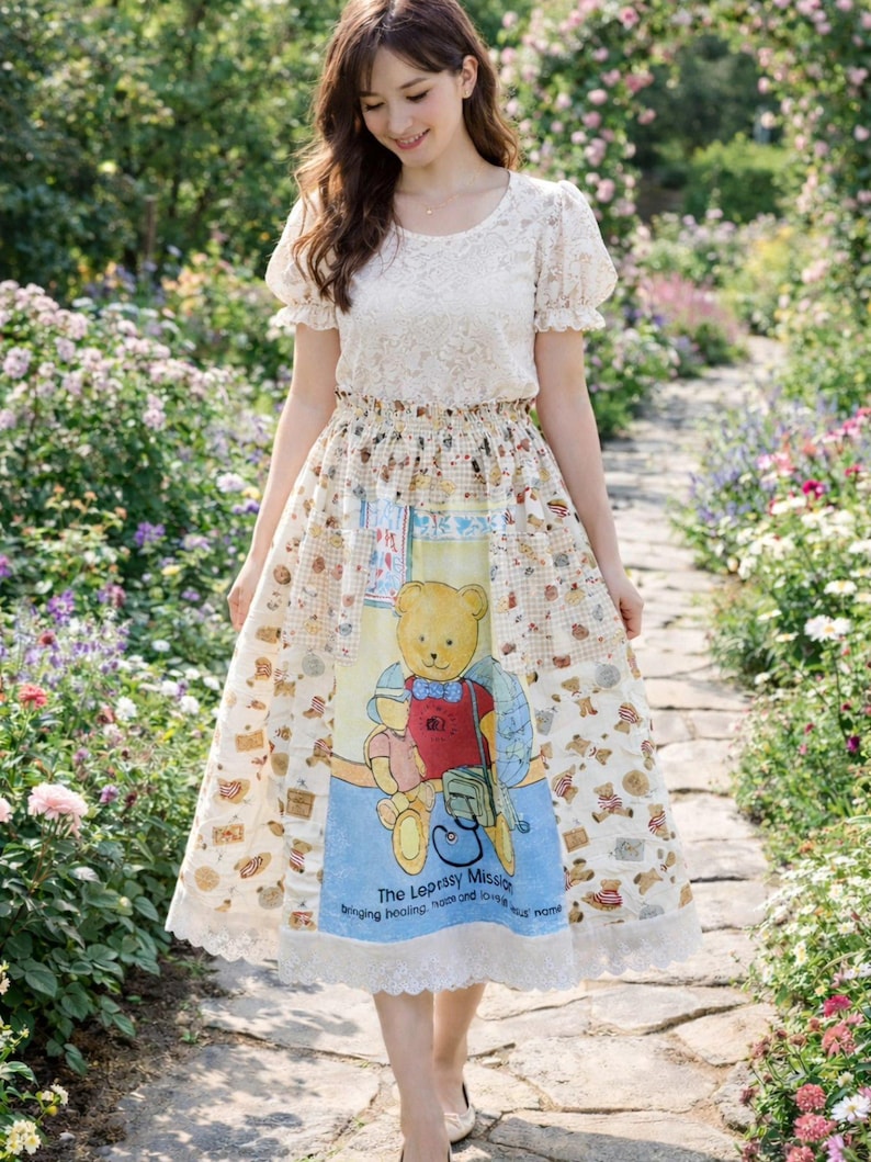 May include: A cream-colored dress with a lace top and a skirt featuring a teddy bear print. The skirt has a blue panel with text and a teddy bear illustration. The dress has a lace trim at the hem.