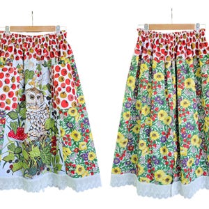 May include: A colorful skirt with a floral and owl print. The skirt features a white background with red berries, yellow flowers, and an owl. The hemline is trimmed with white lace. The skirt is hanging on a wooden hanger.