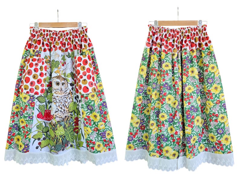 May include: A colorful skirt with a floral and owl print. The skirt features a white background with red berries, yellow flowers, and an owl. The hemline is trimmed with white lace. The skirt is hanging on a wooden hanger.