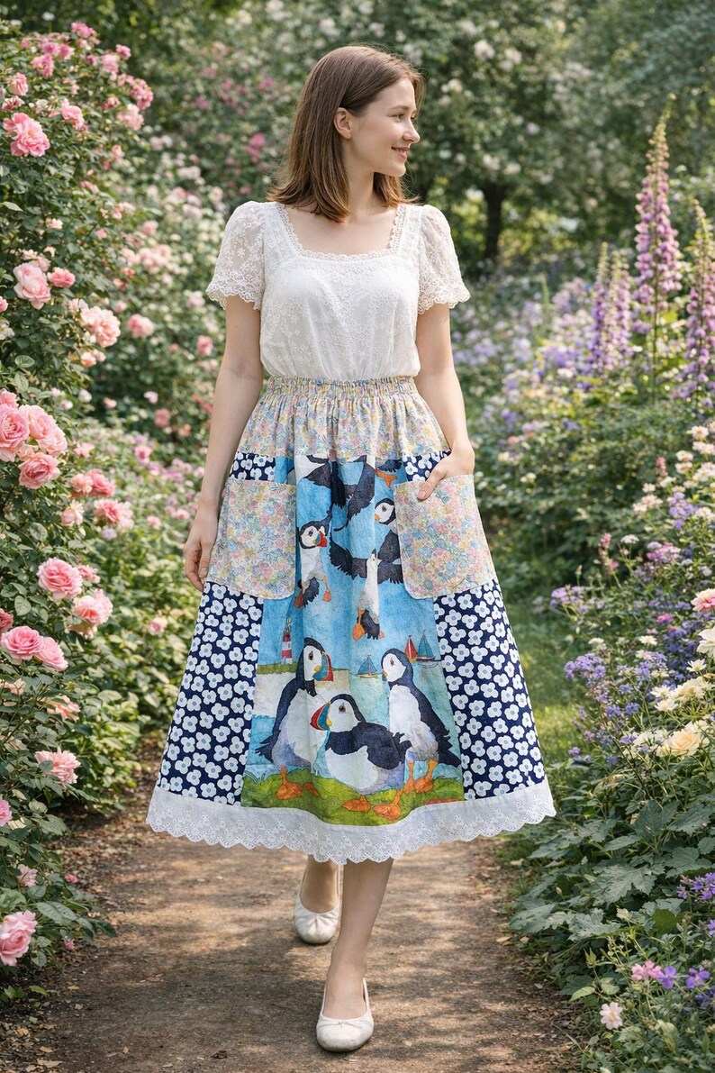 May include: A woman wearing a white lace top and a colorful skirt with a puffin print. The skirt has pockets and a white lace trim. The background features a garden with pink roses and other flowers.