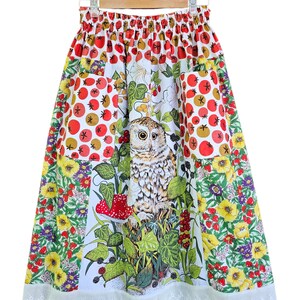May include: A colorful skirt with an owl and floral print. The skirt features a white background with a central panel showcasing an owl surrounded by foliage and a red mushroom. The skirt has pockets and a white lace trim.