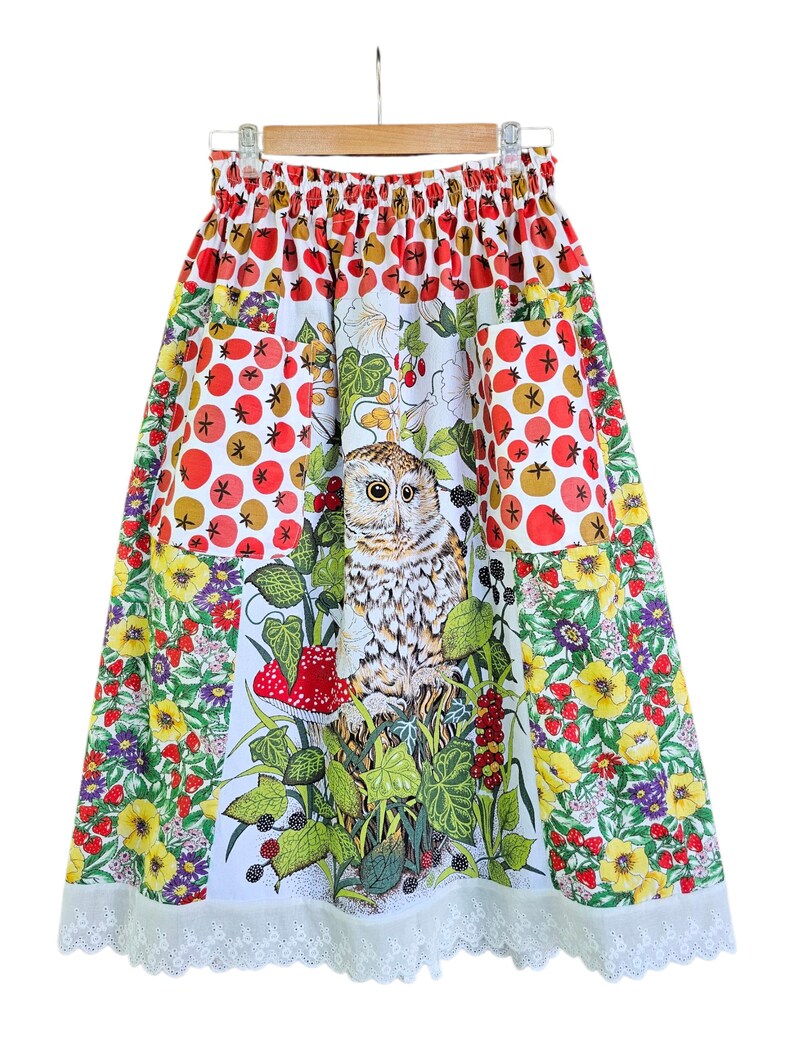 May include: A colorful skirt with an owl and floral print. The skirt features a white background with a central panel showcasing an owl surrounded by foliage and a red mushroom. The skirt has pockets and a white lace trim.