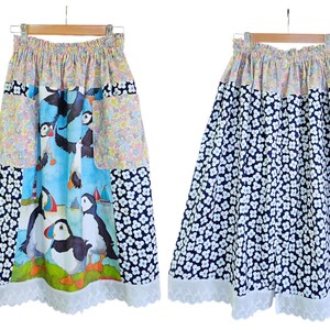 May include: A-line skirt with a colorful puffin print on a blue background, floral patchwork, and a white lace hem. The reverse side features a navy blue floral pattern. The skirt is hanging on a wooden hanger.