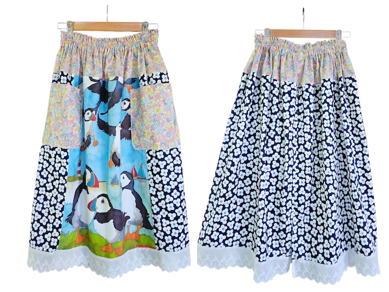 May include: A-line skirt with a colorful puffin print on a blue background, floral patchwork, and a white lace hem. The reverse side features a navy blue floral pattern. The skirt is hanging on a wooden hanger.
