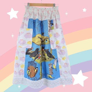 May include: A long, colorful skirt with a birdhouse and bird print. The skirt features a blue center panel with a birdhouse design, flanked by white panels with pink cloud patterns. It has pockets and a lace trim.