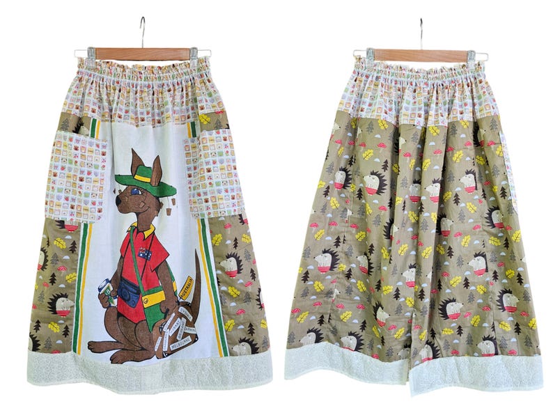 May include: A long skirt with a whimsical design. The front features a kangaroo wearing a hat and carrying luggage. The skirt has a white base with patchwork and a brown and white pattern with hedgehogs. The hem is trimmed with white fabric.