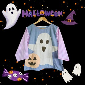 May include: A blue and pink Halloween-themed shirt with a large white ghost and pumpkin applique. The sleeves are pink gingham. The word "Halloween" is displayed above the shirt, along with other Halloween decorations.
