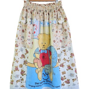 May include: A long, patchwork skirt featuring a large teddy bear graphic on a blue background. The skirt has pockets and a white lace trim. The fabric includes teddy bear and gingham patterns in various colors. Text on the skirt reads "The Leprosy Mission".