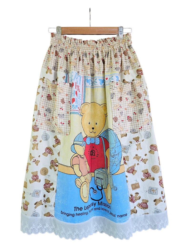 May include: A long, patchwork skirt featuring a large teddy bear graphic on a blue background. The skirt has pockets and a white lace trim. The fabric includes teddy bear and gingham patterns in various colors. Text on the skirt reads "The Leprosy Mission".