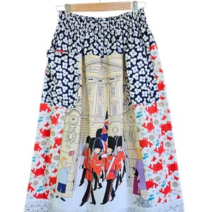 May include: A knee-length skirt with a whimsical design. The top section features a navy blue floral pattern, transitioning to a central panel depicting a building and guards. The lower section has a red crab print and a white, scalloped hem.