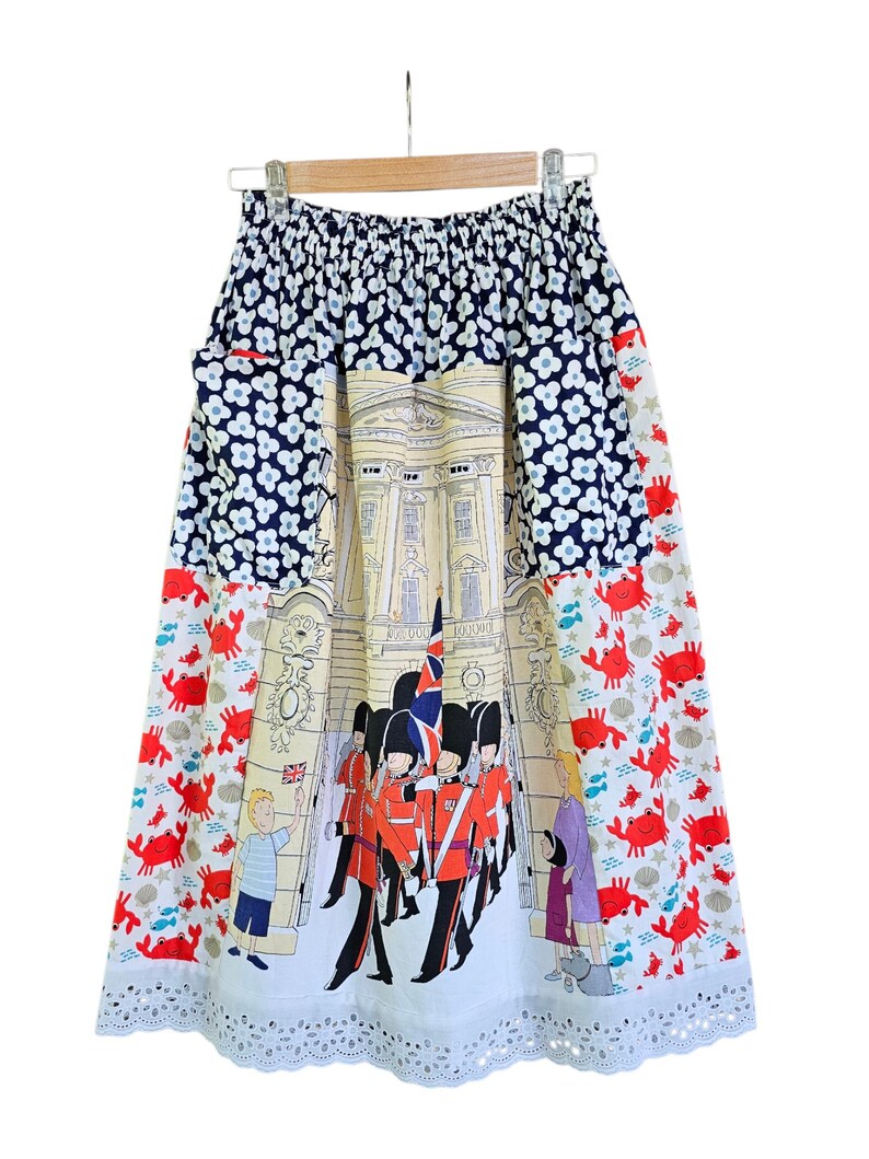 May include: A knee-length skirt with a whimsical design. The top section features a navy blue floral pattern, transitioning to a central panel depicting a building and guards. The lower section has a red crab print and a white, scalloped hem.