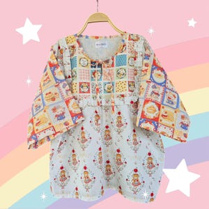 Peut inclure: Une blouse blanche, jaune et bleue en patchwork avec un col à volants et une jupe à imprimé floral. La blouse a un motif de patchwork carré avec une variété de personnages de dessins animés et d'images. La jupe a un fond blanc avec un motif répété de pommes rouges et de filles en robes.