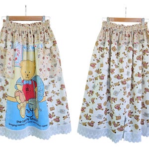 May include: A long, beige skirt with an elastic waistband and a white lace hem. The skirt features a repeating pattern of teddy bears and a central panel with a large teddy bear illustration. The text "The Leapsy Mission" is printed on the skirt.