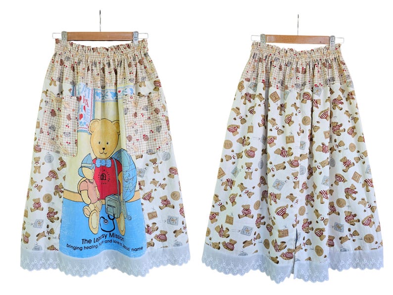 May include: A long, beige skirt with an elastic waistband and a white lace hem. The skirt features a repeating pattern of teddy bears and a central panel with a large teddy bear illustration. The text "The Leapsy Mission" is printed on the skirt.