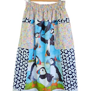 May include: A colorful skirt with a whimsical puffin print. The skirt features a patchwork design with floral and navy blue patterns. The bottom hem is finished with a delicate white lace trim. The skirt is hanging on a wooden hanger.