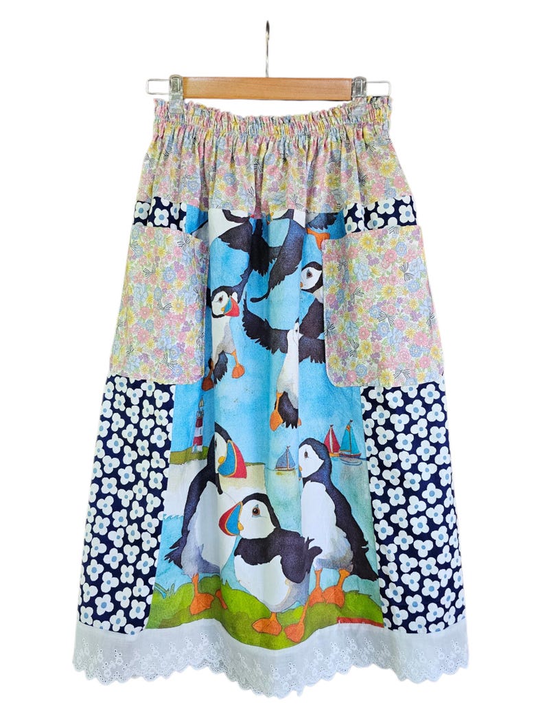 May include: A colorful skirt with a whimsical puffin print. The skirt features a patchwork design with floral and navy blue patterns. The bottom hem is finished with a delicate white lace trim. The skirt is hanging on a wooden hanger.