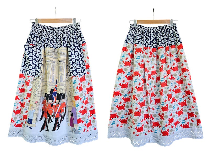 May include: A-line skirt with a unique design. The top section features a navy blue floral pattern, transitioning to a cream-colored panel with a building illustration and red-coated figures. The lower half showcases a white background with red crabs. The skirt has a white eyelet lace hem.
