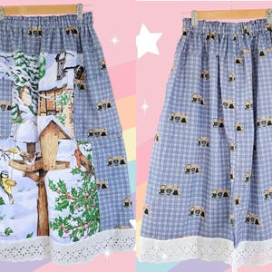 May include: A-line skirt with a blue and white plaid pattern. The skirt features a winter scene with birds, a birdhouse, and holly. The hemline is trimmed with white lace. The skirt is hanging on a wooden hanger.