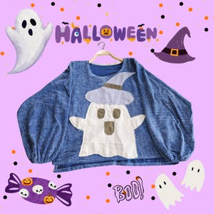Può includere: Una felpa blu a tema Halloween con un'applicazione a forma di fantasma con un cappello da strega. La parola "Halloween" è scritta sopra la maglietta. Ulteriori elementi grafici di Halloween includono fantasmi, caramelle e la parola "Boo!"