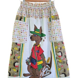 May include: A knee-length skirt with a kangaroo graphic wearing a hat and holding a camera and luggage. The skirt has a white base with a kangaroo illustration, patchwork pockets, and a white lace hem. The skirt is hanging on a hanger.