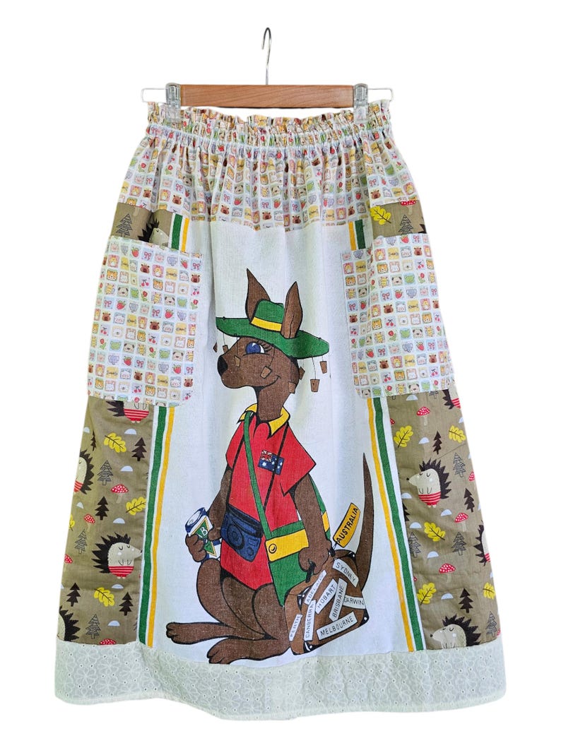 May include: A knee-length skirt with a kangaroo graphic wearing a hat and holding a camera and luggage. The skirt has a white base with a kangaroo illustration, patchwork pockets, and a white lace hem. The skirt is hanging on a hanger.