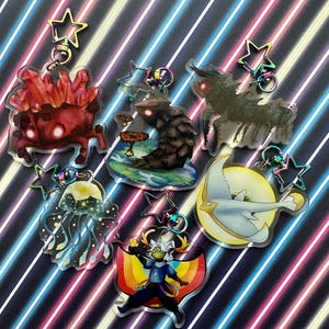 Puede incluir: Seis llaveros de acrílico coloridos con diferentes personajes y diseños. Los llaveros están sobre un fondo a rayas. Los personajes incluyen una criatura roja con ojos brillantes, una criatura marrón con un hongo, una criatura blanca con alas, una medusa azul y una criatura blanca con un arcoíris.