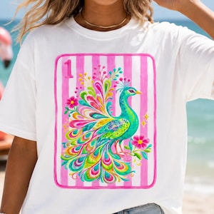 May include: A white short-sleeve t-shirt featuring a rectangular graphic of a colorful peacock with vibrant plumage. The design includes a pink and white striped background, the number 1 in the top left corner, and small pink flowers.