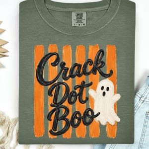May include: An olive green t-shirt featuring a graphic of vertical orange brushstrokes behind the black text "Crack Dot Boo" and a small white ghost illustration. The shirt has a crew neckline and a Comfort Colors brand label at the inner neck.