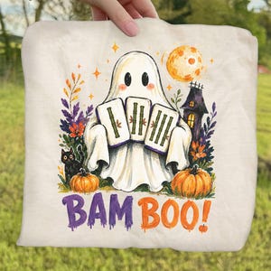 May include: A cream-colored fabric item featuring a graphic of a white ghost holding three bamboo tiles. The design includes a black cat, orange pumpkins, a dark house, and a full moon. The text BAM BOO! is printed in purple and orange below.