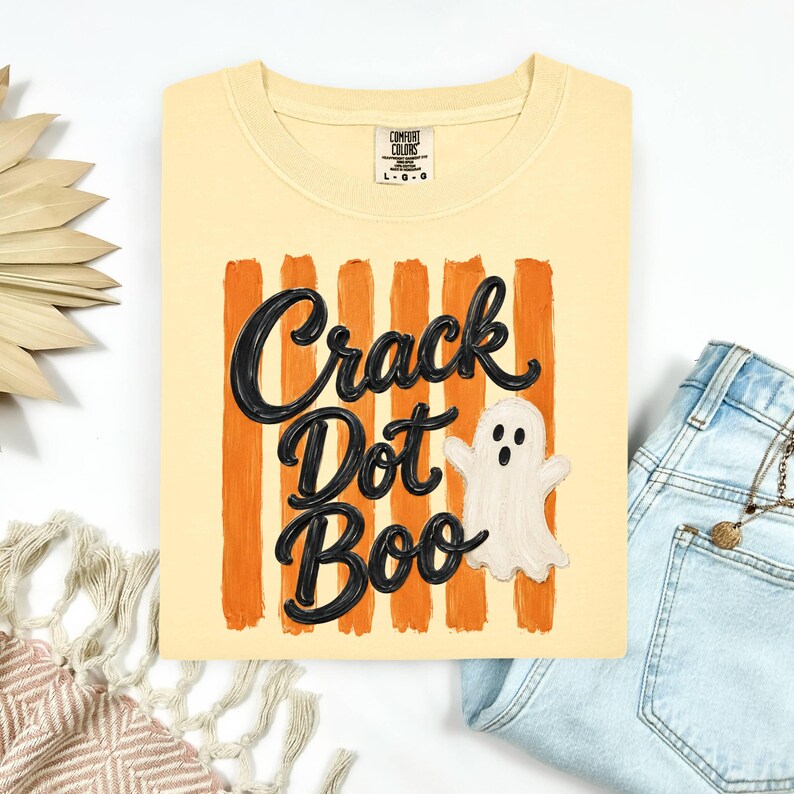 May include: A pale yellow t-shirt featuring a graphic of vertical orange brushstrokes, the black text "Crack Dot Boo" in a cursive font, and a small white ghost illustration. The shirt has a Comfort Colors brand label at the inner neckline.