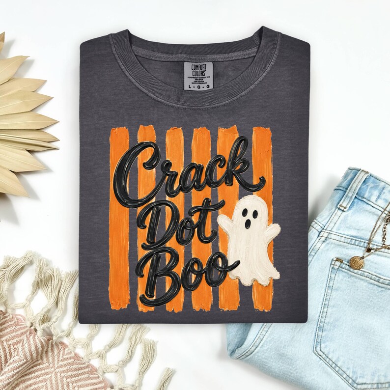 May include: A charcoal gray t-shirt featuring a graphic of vertical orange brushstrokes behind the black text "Crack Dot Boo" and a white ghost illustration. The shirt has a crew neckline and a Comfort Colors brand label at the inner collar.