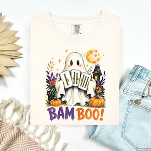 May include: A cream-colored t-shirt featuring a graphic of a ghost holding three bamboo tiles, a black cat, pumpkins, a haunted house, and a full moon. The text BAM BOO! is printed in purple and orange below the illustration. The shirt has a Comfort Colors label.