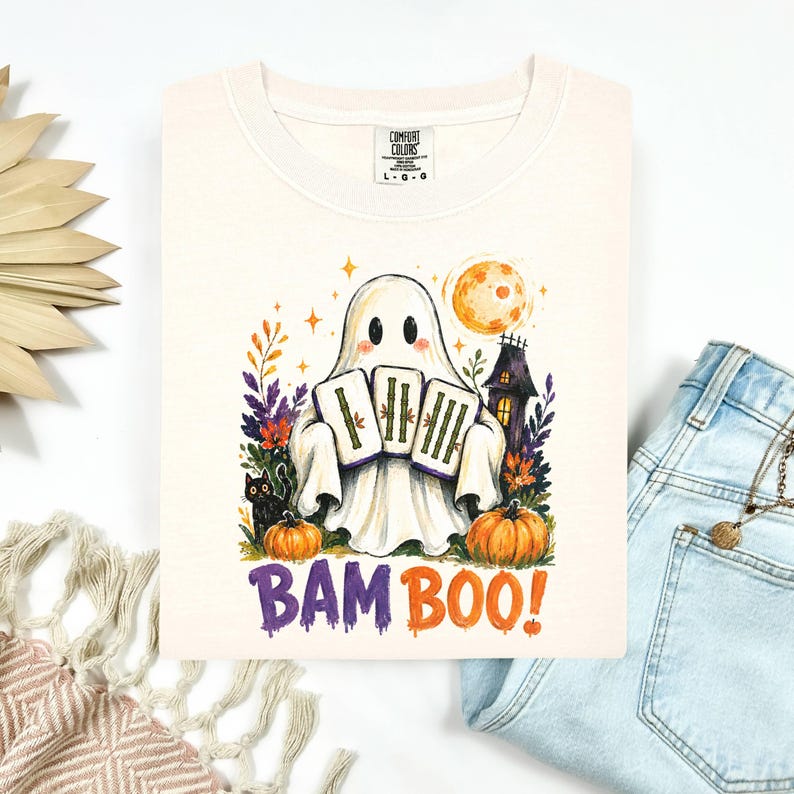 May include: A cream-colored t-shirt featuring a graphic of a ghost holding three bamboo tiles, a black cat, pumpkins, a haunted house, and a full moon. The text BAM BOO! is printed in purple and orange below the illustration. The shirt has a Comfort Colors label.